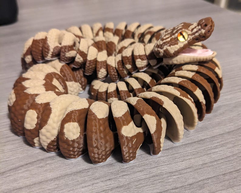 May include: A 3D-printed articulated snake toy in shades of brown and cream. The snake's body is segmented with a repeating pattern. The head is detailed with a realistic eye and open mouth, showcasing a pink interior.