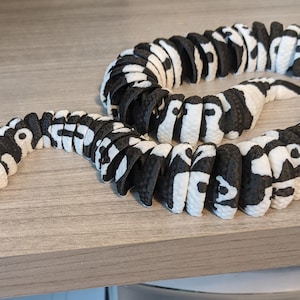 May include: A black and white toy snake with a patterned design. The snake is made of connected segments, with a white head and black and white body. The toy is lying on a light gray surface.