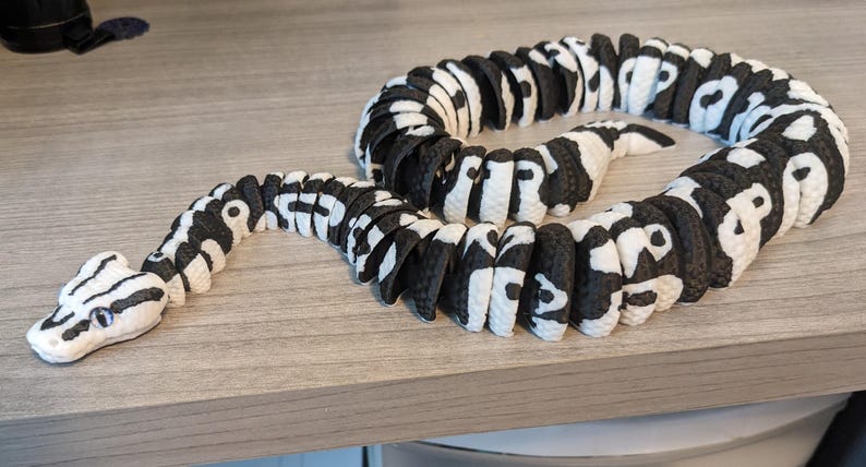 May include: A black and white toy snake with a patterned design. The snake is made of connected segments, with a white head and black and white body. The toy is lying on a light gray surface.