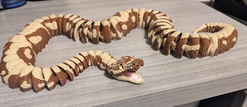 May include: A segmented toy snake in shades of brown and cream, with a detailed head and open mouth. The snake is posed on a light gray surface, showcasing its articulated body.