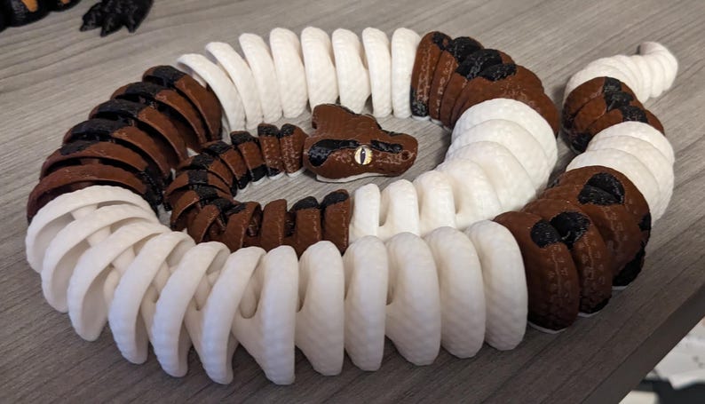 May include: A 3D-printed snake toy in a coiled position. The snake features alternating segments of white and brown with black spots. The head is brown with a yellow eye. The toy is made of plastic and sits on a wooden surface.