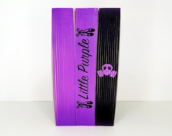 May include: Three decorative books with purple and black covers. The purple book has the text "Little Purple" with ballet shoes. The black book has a gas mask symbol. The books are standing upright.
