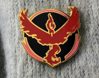 May include: A gold-colored pin with a red and black design. The design features a stylized bird with a flame-like crest, surrounded by a pink circle.
