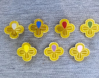 May include: Seven gold-colored clover-shaped enamel pins with different colored center accents: white, blue, red, green, yellow, purple, and pink. The pins are on a gray textured surface.