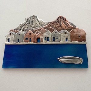 May include: A metal artwork showing a coastal village scene. It features a row of small houses with different colours and textures, set against mountains and a blue body of water. A small boat is also present in the scene.