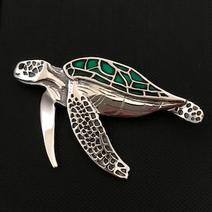 May include: A silver sea turtle pendant with a green enamel shell. The turtle has detailed sculpting and a textured shell.