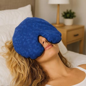 May include: A blue eye mask with a black trim covers the eyes of a person lying on a bed. The mask is made of a textured fabric. The person is wearing a white tank top. A lamp and nightstand are in the background.