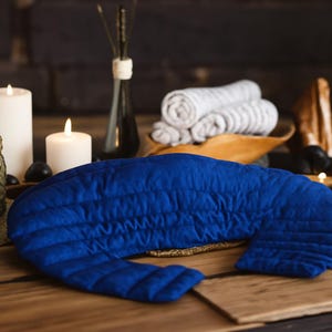 May include: A vibrant blue, quilted neck wrap rests on a wooden surface. The wrap is surrounded by lit candles, rolled towels, and decorative items, creating a spa-like ambiance. The wrap's design suggests it is for relaxation and comfort.