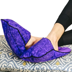 May include: A pair of purple foot warmers with a marbled pattern and black trim. The foot warmers are placed on a patterned pillow. The person is wearing black leggings.