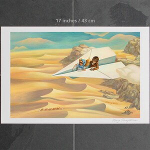 May include: A framed art print, 17 x 11 inches, depicts two figures riding a paper airplane over a desert landscape. The artwork features warm tones of yellow and brown, with a blue sky and clouds. A caravan of camels is visible in the distance.