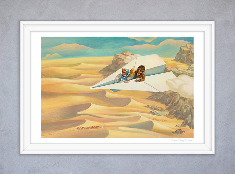 May include: A framed art print depicts two figures in aviator gear riding a paper airplane over a desert landscape. The artwork features warm tones of yellow and brown, with a blue sky and clouds. The print is signed.