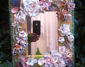 May include: A decorative mirror with a frame made of seashells and other natural materials. The frame is in shades of white, pink, and green. The mirror is surrounded by a variety of flowers, including roses, daisies, and lilies.