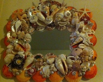 May include: A mirror with a frame made of seashells. The frame is made of a variety of seashells in different shapes and sizes. The seashells are arranged in a circular pattern around the mirror. The mirror is surrounded by a border of orange seashells.