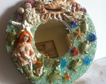 May include: A round decorative mirror with a mermaid and sea life theme. The mirror is surrounded by seashells, coral, starfish, and small turtles. The mermaid has red hair and is holding a starfish. The frame is a light green color.