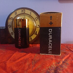 May include: A black Duracell battery with a gold top and the Duracell logo, next to its original box. The box is brown and black with the Duracell logo and an arrow pointing up. A vintage clock is in the background.