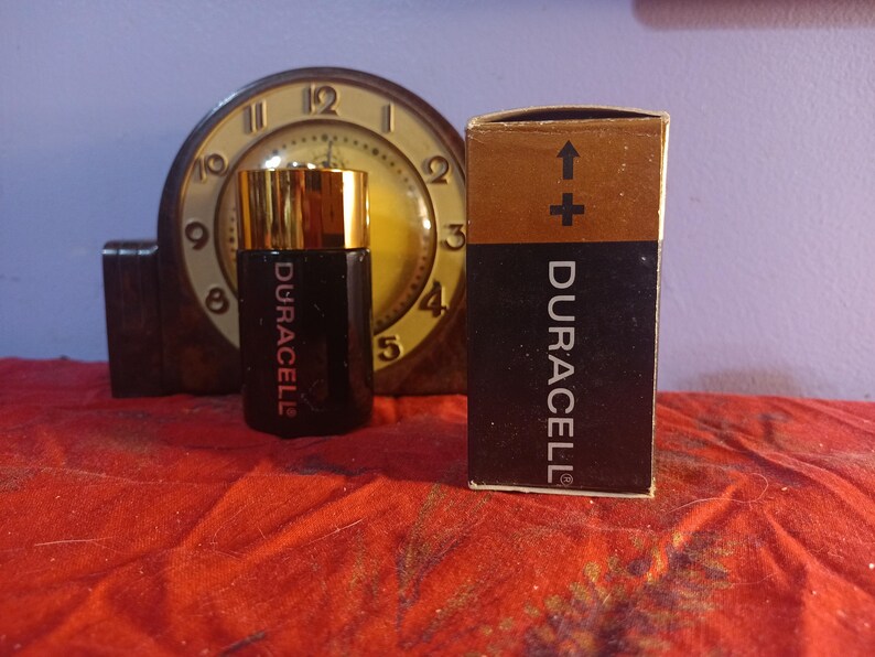 May include: A black Duracell battery with a gold top and the Duracell logo, next to its original box. The box is brown and black with the Duracell logo and an arrow pointing up. A vintage clock is in the background.