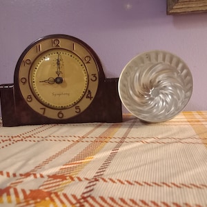 May include: A vintage brown clock with a gold face and the word "Symphony" printed on it. Next to the clock is a silver, spiral-shaped baking mold. The clock shows the time as approximately 1:00.