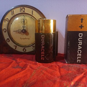 May include: A vintage clock with a gold-coloured face and black hands, next to a Duracell battery and its box. The battery is black with gold accents and the Duracell logo. The box is black and gold with the Duracell logo.