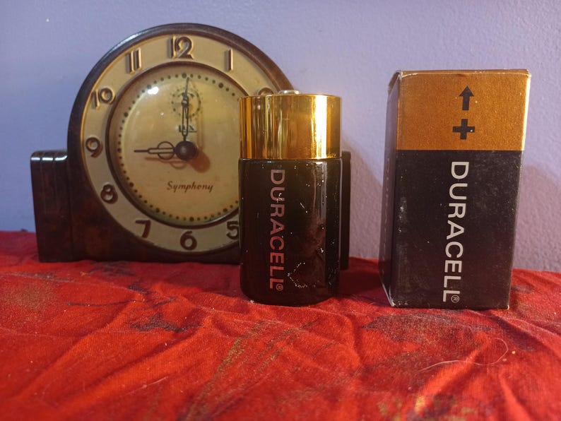 May include: A vintage clock with a gold-coloured face and black hands, next to a Duracell battery and its box. The battery is black with gold accents and the Duracell logo. The box is black and gold with the Duracell logo.
