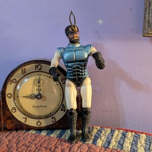 May include: A plastic action figure with a blue chest piece, black gloves, black boots, and a gold antenna on its head, standing in front of a brown wooden mantle clock with a cream-colored face that displays the text Symphony and U.S.