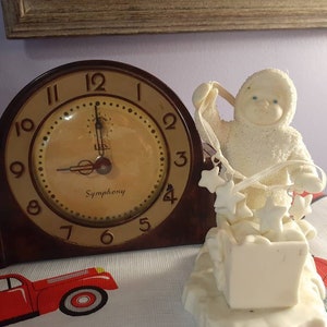 May include: A white porcelain figurine of a snowman holding a string of stars. The snowman is wearing a hat and scarf and is sitting on a base with a box. A brown clock with a white face and black hands is in the background.
