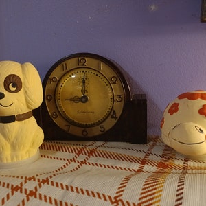 May include: A collection of vintage ceramic figurines and a clock. The figurines include a dog with brown accents and a turtle with orange flower details. The clock has a dark brown frame and a gold-colored face with the word "Symphony".