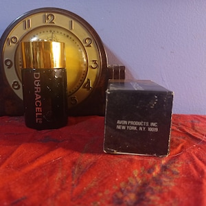 May include: A vintage black perfume bottle with a gold cap, labelled "Duracell", is positioned in front of a classic clock. A black box with the text "AVON PRODUCTS, INC NEW YORK, NY 10019" is also present. The items are on a red surface.