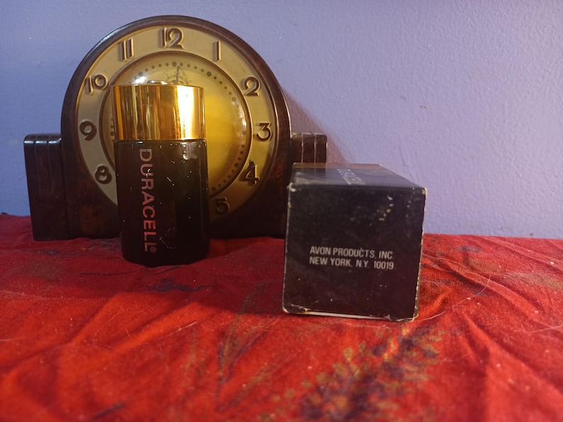 May include: A vintage black perfume bottle with a gold cap, labelled "Duracell", is positioned in front of a classic clock. A black box with the text "AVON PRODUCTS, INC NEW YORK, NY 10019" is also present. The items are on a red surface.