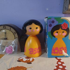 May include: A vintage yellow doll with a black hair and pink floral design. The doll is next to its original box with a matching illustration. A clock and a small bag with a pink flower are also in the image.