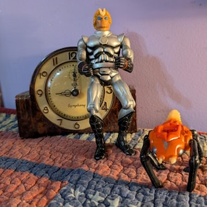 May include: A standing action figure with blonde hair, blue eyes, and silver armor, positioned next to a small orange and black spider-like creature. Behind them is a wooden mantle clock with a cream-colored face and the word Symphony printed on it.