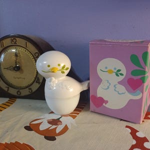 May include: A white dove-shaped container with a yellow beak and green leaves, next to a matching box with a dove illustration and a pink heart. A vintage clock with a brown wooden frame is in the background.