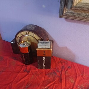 May include: A vintage clock with a brown face and gold hands is behind a black and orange Duracell battery and a box of Duracell batteries. The box has the text "SUPER CHARGE" and "1.5 FL OZ".