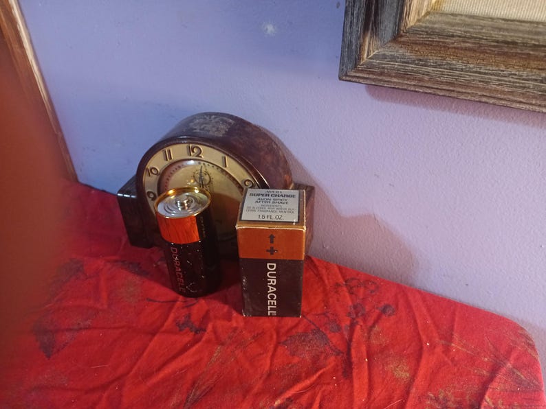 May include: A vintage clock with a brown face and gold hands is behind a black and orange Duracell battery and a box of Duracell batteries. The box has the text "SUPER CHARGE" and "1.5 FL OZ".