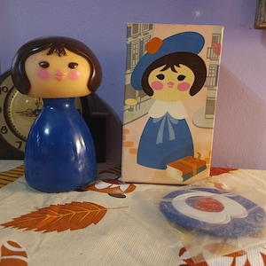 May include: A vintage blue plastic doll with a brown hair and a round face, next to a box with a drawing of a girl in a blue dress and hat. A blue and white circular object is in a plastic bag.
