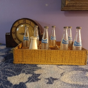 May include: A vintage wooden crate holds six small glass bottles of Fanta soda, each with a blue and white label. A white ceramic salt shaker sits in the crate. An antique clock is in the background.