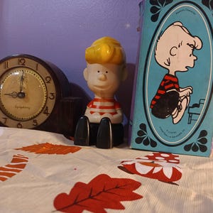 May include: A vintage clock, a figurine of a cartoon character, and a book with a cartoon illustration are displayed on a patterned surface. The clock has a dark brown frame and a cream-colored face. The figurine is wearing a red and white striped shirt. The book has a blue cover.