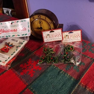 May include: Christmas craft kits and holly sprigs are displayed on a red and green plaid surface. The kits feature reindeer and the text "Christmas Kits." Two packages of holly with red berries are labeled "Home For The Holidays."