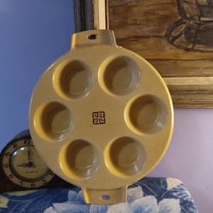 May include: A beige ceramic baking pan with six round molds. The pan has a handle on each side and a decorative logo in the center.