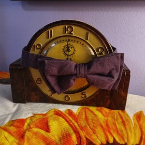 May include: A vintage brown Bakelite clock with a gold face and black Roman numeral markings. A purple bow tie is tied around the clock face.