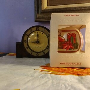 May include: A white ceramic Christmas ornament with a red poinsettia and a basket of oranges and a wrapped gift. The ornament is in a clear plastic box with the words "Grandparents" and "Keepsake Ornament" printed on the box.