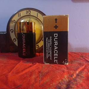 May include: A black Duracell battery and its box are displayed on a red surface, with a vintage clock in the background. The box has a gold top and the Duracell logo. The battery is black with gold accents and the Duracell logo.