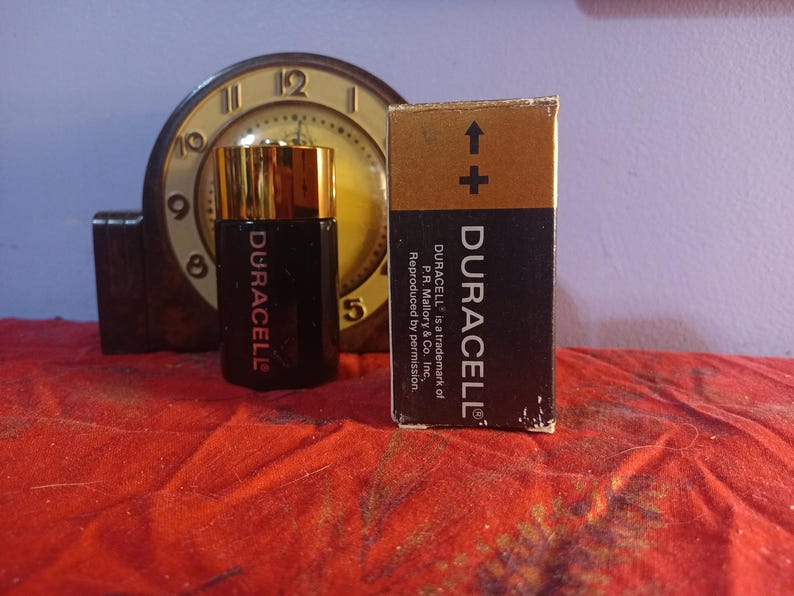 May include: A black Duracell battery and its box are displayed on a red surface, with a vintage clock in the background. The box has a gold top and the Duracell logo. The battery is black with gold accents and the Duracell logo.