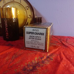 May include: An Avon Super Charge aftershave box and bottle with a gold cap. The box reads "Avon Spicy After Shave" and "1.5 fl oz." The bottle is dark with the word "Duracell" on it. A vintage clock is in the background.
