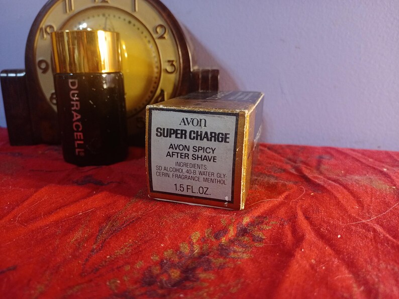 May include: An Avon Super Charge aftershave box and bottle with a gold cap. The box reads "Avon Spicy After Shave" and "1.5 fl oz." The bottle is dark with the word "Duracell" on it. A vintage clock is in the background.