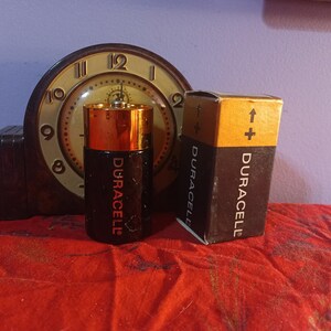 May include: A Duracell D-cell battery and its packaging are displayed in front of a vintage clock. The battery is black and gold, with the Duracell logo in red. The packaging is brown and black, with the Duracell logo and a plus sign.