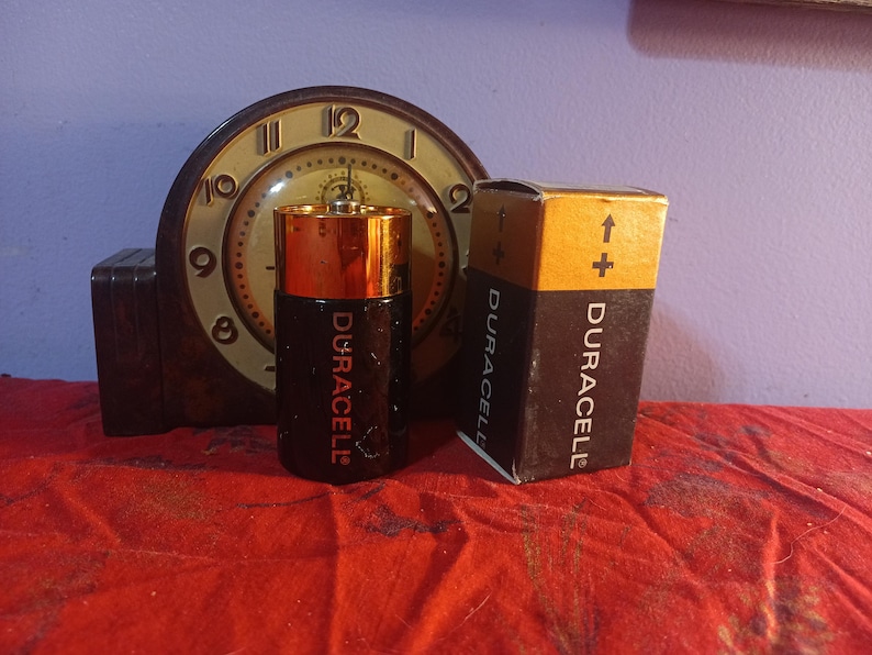 May include: A Duracell D-cell battery and its packaging are displayed in front of a vintage clock. The battery is black and gold, with the Duracell logo in red. The packaging is brown and black, with the Duracell logo and a plus sign.