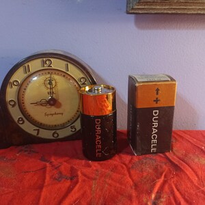 May include: A vintage clock with a brown wooden case and a cream-coloured face, beside a Duracell battery and its box. The battery is black and gold, and the box is brown and black. The clock face reads "Symphony".