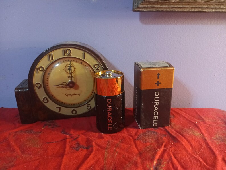 May include: A vintage clock with a brown wooden case and a cream-coloured face, beside a Duracell battery and its box. The battery is black and gold, and the box is brown and black. The clock face reads "Symphony".