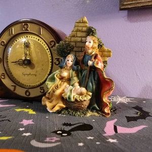 May include: A ceramic nativity scene figurine set featuring Mary, Joseph, and baby Jesus. The figures are painted in realistic colors and are posed in a traditional nativity scene setting. The figurine set is displayed on a table with a gray and white patterned tablecloth.