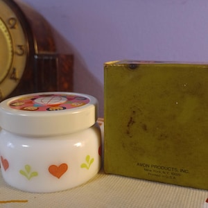 May include: A white jar with a decorative lid featuring colorful cartoon faces. The jar has orange hearts and green leaf designs. Next to it is a green box with text that reads "AVON PRODUCTS, INC. New York, N.Y. 10020 Printed in U.S.A."